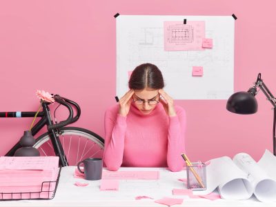 Serious talented young European female architect being busy at her workplace makes project of new residential complex concentrated in papers wears casual turtleneck tries to gather with thoughts